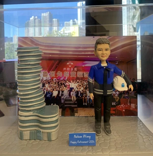 一款為興聯集團的資深工程師Nelson Wong訂製的退休禮物人像公仔，被封存在透明展示盒中。公仔身穿工程制服、手持安全帽，自信地站在一座獨特的建築模型旁，背景是公司團隊在「魯班先師寶誕」活動上的大合照，象徵著對其畢生貢獻的最高敬意。