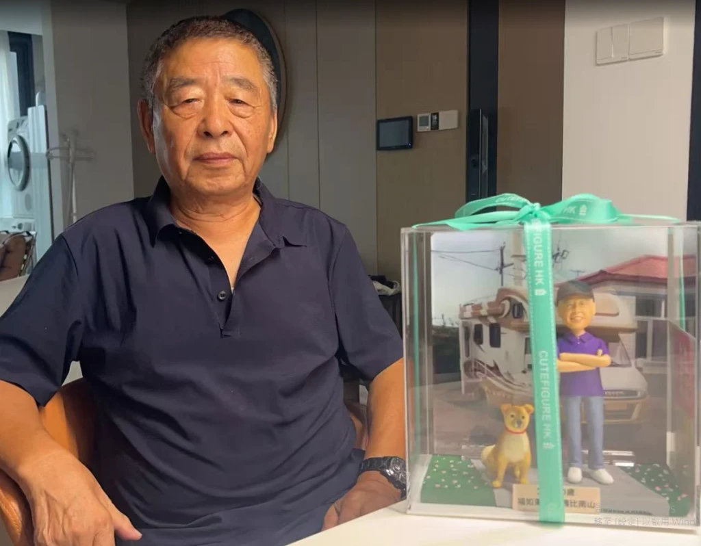 這是一張真人與人像公仔對比圖，主角坐在桌旁，桌上擺著一份精緻的人像公仔退休禮物。禮物盒內有一個微笑的人像公仔和一隻小狗，背景照片展示了一台露營車和房屋，細緻還原退休人士的生活風景。