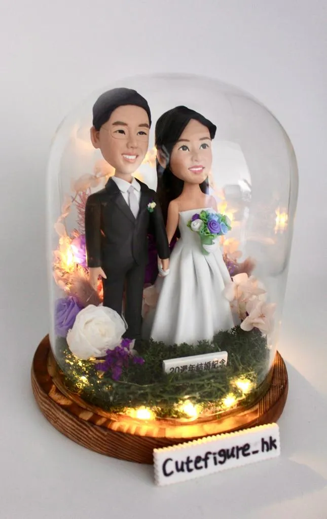 一對新人婚紗公仔，男方西裝、女方白紗，手捧花束，站在玻璃罩內，底部有燈飾和花卉裝飾。