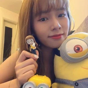 一位可愛的年輕女性，與她的Minions（迷你兵團）收藏品合照，她手中舉著一個根據她樣貌訂製的人像公仔，公仔還抱著一隻白色小兔子，是一款充滿童趣的個人化禮物。