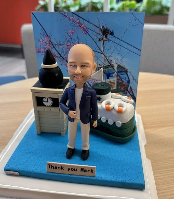 一個穿著藍色外套和米色褲子的人像公仔站在立體場景中，背景有櫻花、電線杆和輪船元素，旁邊有時鐘塔和感謝字牌，色彩鮮明。