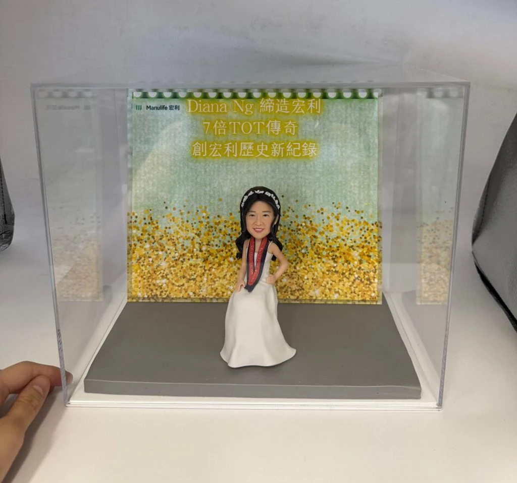 一款女性人像公仔，穿著白色禮服並佩戴獎牌，坐姿展示自信笑容，背景有金色閃爍裝飾和祝賀語，內容包括「締造宏利7倍TOT傳奇」等。這款人像公仔是專為升職禮物、上司禮物、公司禮物及業職祝賀禮物而設計，突顯職業成就。