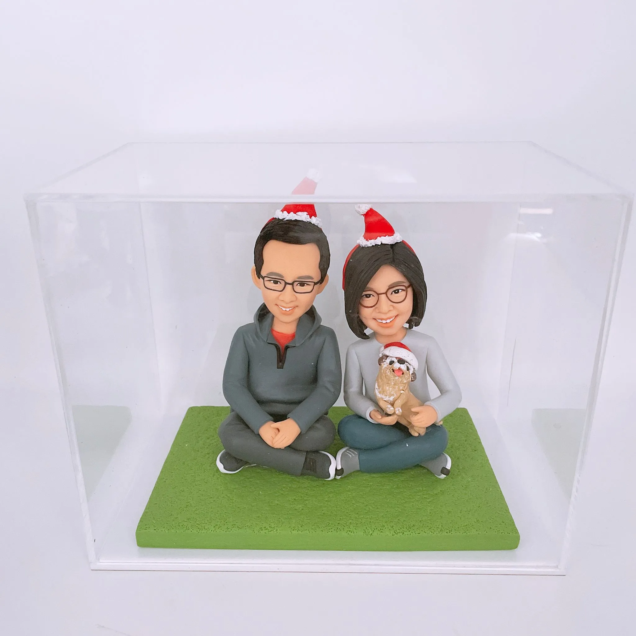 一對情侶和一隻狗的人像公仔，頭戴聖誕帽，坐在綠色草地造型底座上，裝在透明盒內。