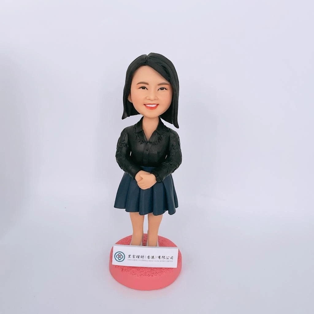 一隻單人像公仔，女性造型，身穿黑色襯衫和深藍色裙子，雙手自然交疊於身前，站在粉紅色圓形底座上，底座前方有公司名牌。這款人像公仔適合作為升職禮物，強調單人像公仔的設計特色。
