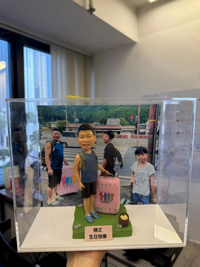 一個穿著休閒服裝、背著背包並拉著行李箱的小朋友人像公仔，放在透明展示盒內，背景是與家人在戶外的合照，底座有祝福字牌。