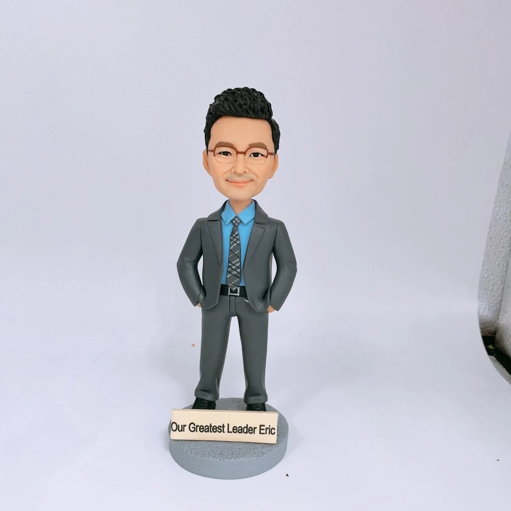 一隻單人像公仔，穿著灰色西裝、藍色恤衫和花紋領帶，雙手插袋，站在圓形底座上，底座前方有「Our Greatest Leader Eric」字牌。這款人像公仔適合作為升職禮物，突顯單人像公仔的設計特色。