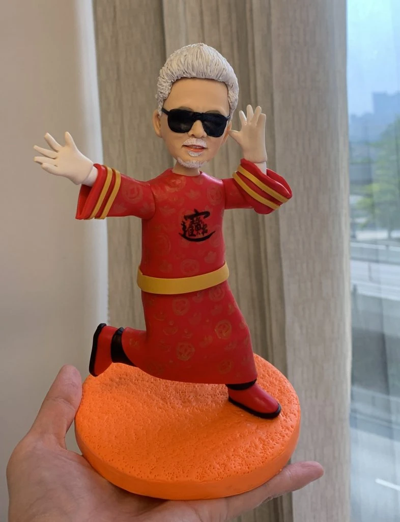 一隻男士人像公仔，身穿紅色傳統服飾並配有金色飾邊，頭髮白色，戴著黑色太陽眼鏡，擺出幽默搞笑動作，單腳站在橙色圓形底座上。這款人像公仔以幽默、搞笑造型為主題，適合用作升職禮物。