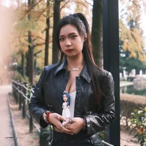 一位穿著黑色皮衣、梳著高馬尾的時尚女性，在秋日的公園裡，雙手捧著一個與她風格形成對比的人像公仔，公仔有著藍色頭髮並穿著白色長裙，是一款展現個人多面性的獨特禮物。
