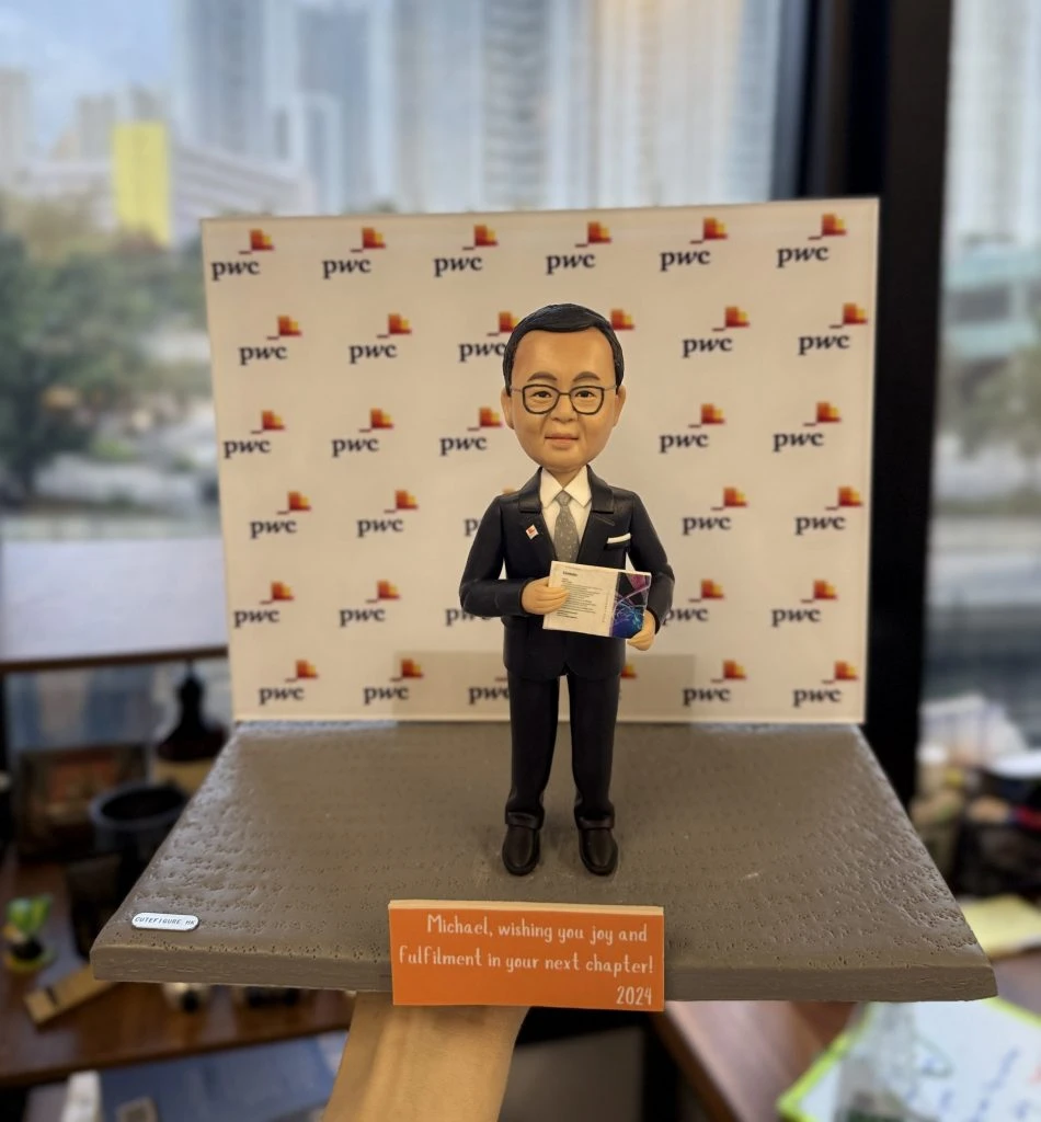 一款男性人像公仔，穿著西裝並手持文件，站立於灰色平台上，身後是印有公司LOGO（pwc）的背景牆。這款人像公仔非常適合作為升職禮物、上司禮物及公司禮物，突顯專業形象與企業歸屬感。