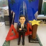 一款訂製的人像公仔升職禮物，適合作為上司禮物或公司禮物。公仔穿著西裝，站立於灰色磚紋地板上，背後有紅色立體公司AON字母和一座黃色星形獎盃，上面寫有「Best Boss Award」和「#Team6」。背景為太古坊港島東中心高樓的景觀照片，突顯專業與榮耀。