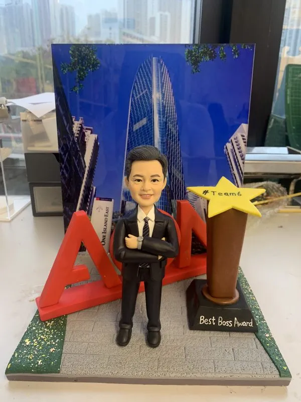 一款訂製的人像公仔升職禮物，適合作為上司禮物或公司禮物。公仔穿著西裝，站立於灰色磚紋地板上，背後有紅色立體公司AON字母和一座黃色星形獎盃，上面寫有「Best Boss Award」和「#Team6」。背景為太古坊港島東中心高樓的景觀照片，突顯專業與榮耀。