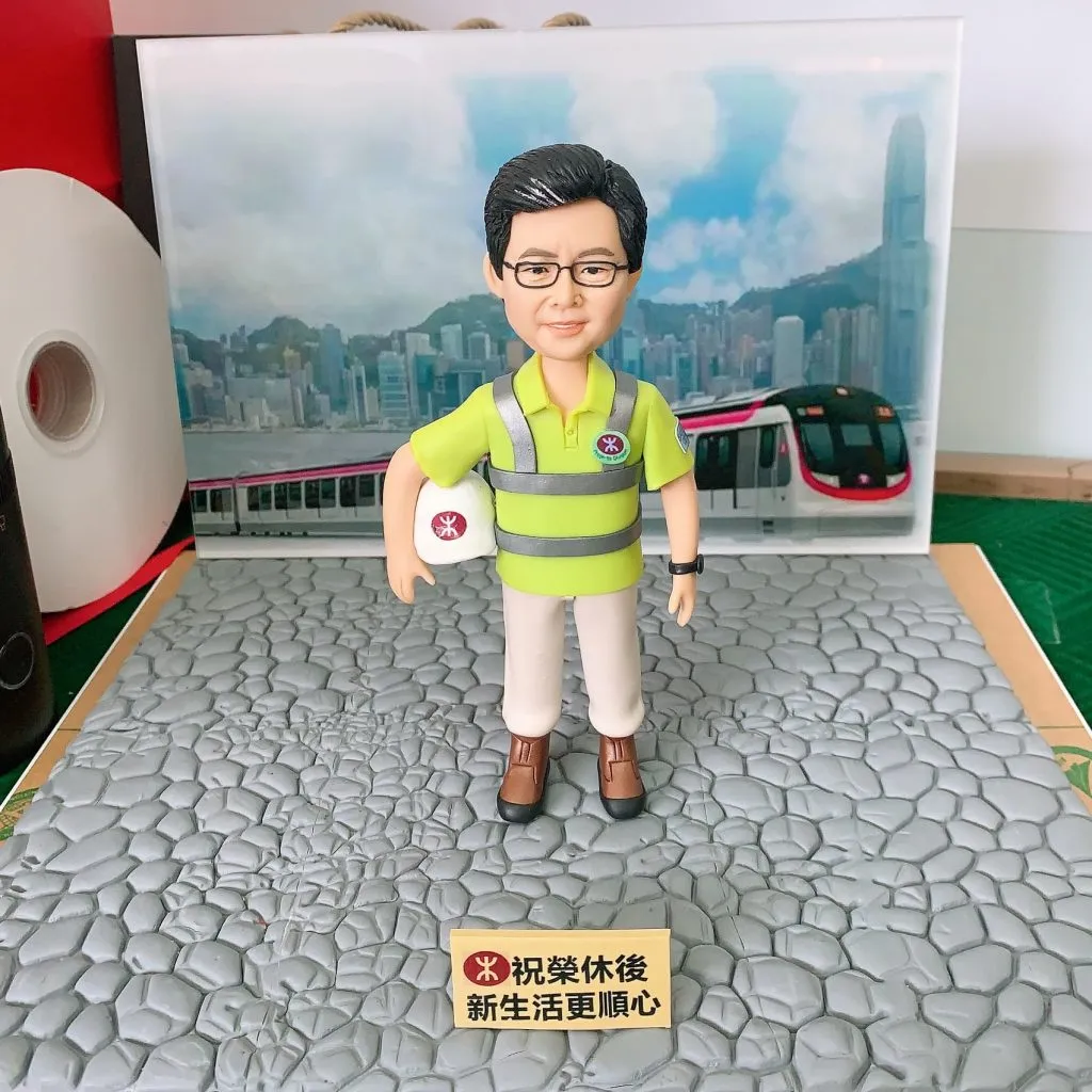 一位港鐵員工的人像公仔，他身穿反光衣制服、手持安全帽，背景是香港維多利亞港和港鐵列車。這是一款送給同事的榮休禮物，底座銘牌上寫有祝福語，祝願他退休後生活順心。