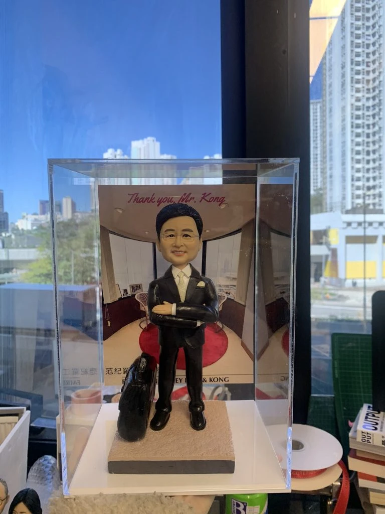 一款專為升職或表揚上司設計的人像公仔公司禮物，公仔穿著西裝，雙手交叉站立，腳邊放有黑色公事包，背景為辦公室場景，