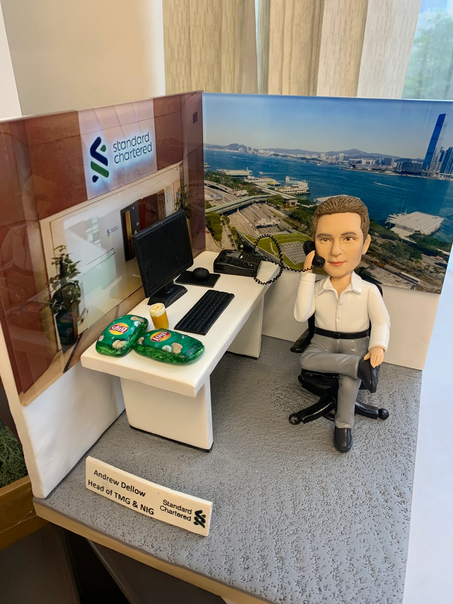 男士人像公仔穿著白色恤衫和灰色西褲，坐在辦公椅上用電話交談。前方有辦公桌、電腦和零食。立體背景包括公司門口標誌以及公司窗外的海景城市景色。這款人像公仔適合作為升職禮物、上司禮物或公司禮物，特色是立體背景、公司門口及窗外景色。