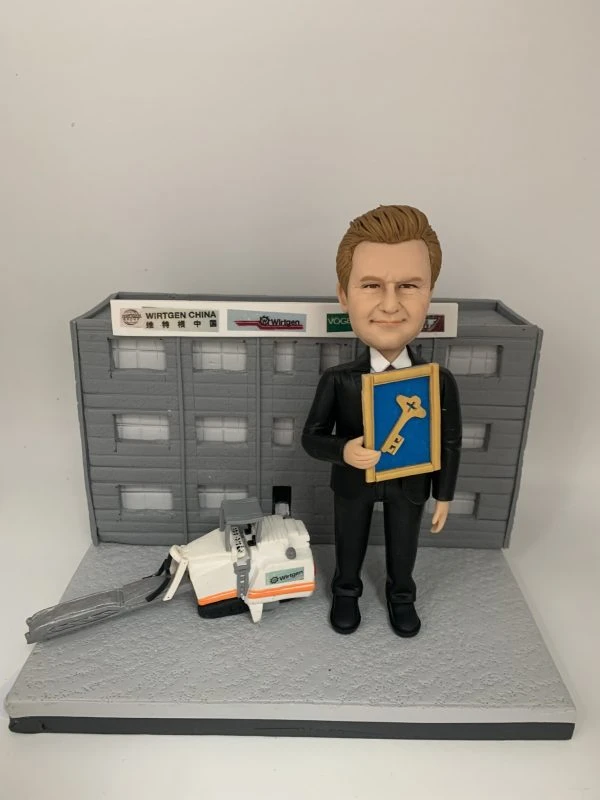 男士人像公仔身穿西裝，手持獎狀，站在立體建築背景前，旁邊配有立體工程車模型。背景建築牆上有公司標誌，整體設計突出立體背景、立體建築和工程車元素。這款人像公仔適合作為升職禮物、上司禮物或公司禮物。