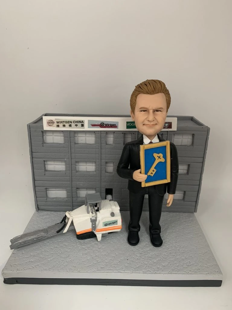 男士人像公仔身穿西裝，手持獎狀，站在立體建築背景前，旁邊配有立體工程車模型。背景建築牆上有公司標誌，整體設計突出立體背景、立體建築和工程車元素。這款人像公仔適合作為升職禮物、上司禮物或公司禮物。