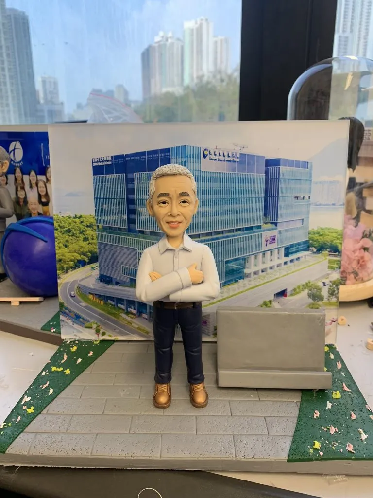 一個銀髮男士的人像公仔卡片座，公仔雙手抱胸，充滿自信地站在一座宏偉的現代建築照片前，此為一款客製化的升職禮物或工程師禮物，用以表彰其專業成就。