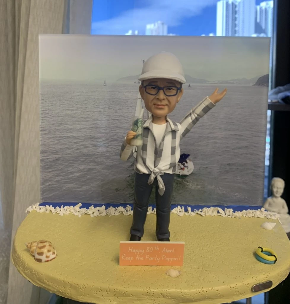 一個戴著白帽和眼鏡、穿著休閒襯衫的人像公仔站在沙灘風格的底座上，手持飲品並舉手慶祝，背景是大海和帆船，底座有80大壽祝福字牌。