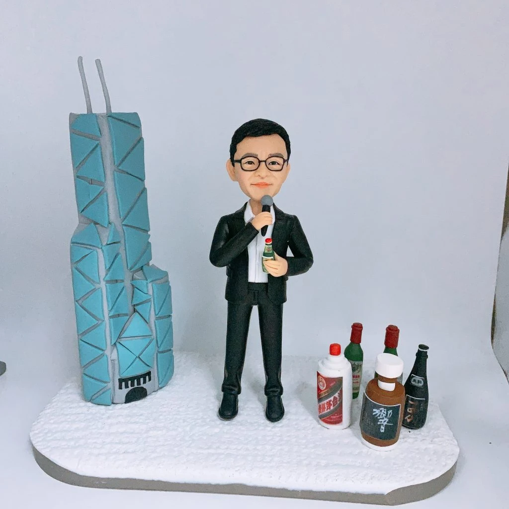 男士人像公仔身穿西裝，手持麥克風和酒瓶，站在白色底座上。左側有立體辨識度高的建築模型，右側擺放多款立體酒瓶。這款人像公仔適合作為升職禮物、上司禮物或公司禮物，特色是立體背景、立辨建築和酒元素。