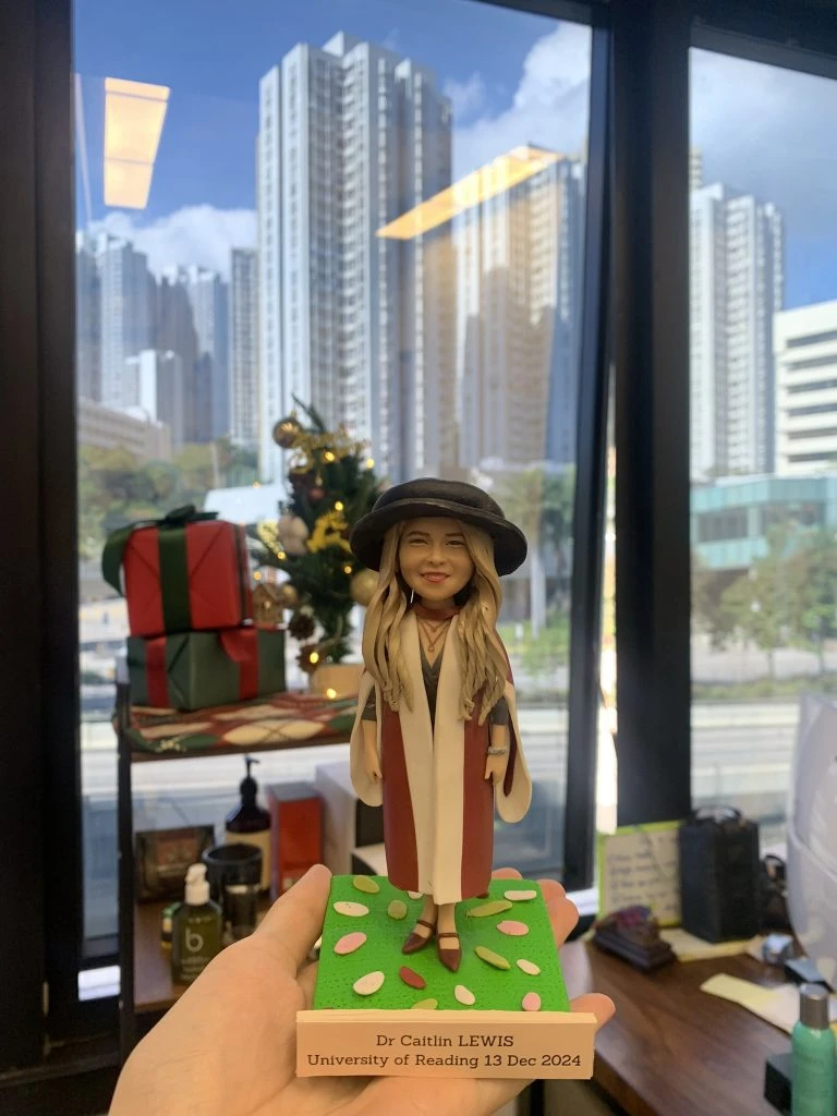 一個女性人像公仔，身穿英式博士畢業袍，外披紅白相間的長袍，內穿深色服裝，頭戴黑色博士帽，長髮微捲，面帶笑容，公仔底座寫有「Dr Caitlin LEWIS University of Reading 13 Dec 2024」。