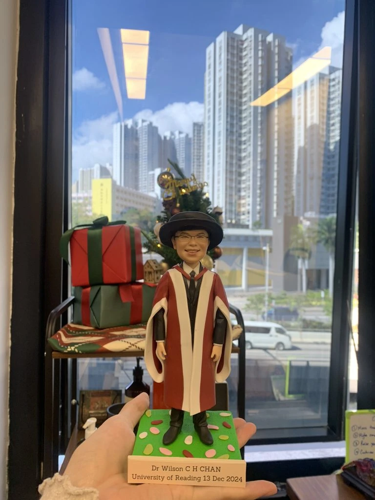 一個男人人像公仔，穿著雷丁大學的紅白博士畢業袍和黑色學位帽，戴著眼鏡，手持畢業證書，站在寫有「Dr Wilson C H CHAN University of Reading 13 Dec 2024」的底座上。