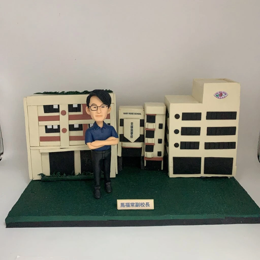 天保民學校馬福常副校長的人像公仔，他雙臂交叉，自信地站立在精細的學校建築模型前。這是一款極具份量的榮休禮物，象徵著他對學校的貢獻。