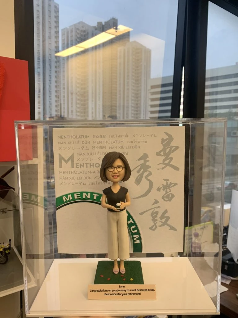 一款專為升職或退休定制的人像公仔，公司禮物或上司禮物首選。公仔是一位女性，手持公司產品，站在綠色草地上，背景為公司品牌曼秀霤敦的大型LOGO與多語言公司名稱