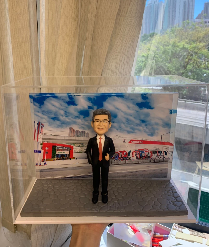 一個以男性為主角的人像公仔，身穿黑色西裝和紅色領帶，站立在灰色磚紋平台上，背景是公司加油站（SINOPEC）和藍天白雲的公司外景。這款公仔適合作為升職禮物、上司禮物或公司禮物，強調公司背景元素。