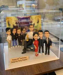 八位人像公仔站在一起合照，中央一位男士坐在車上，手持飲品，旁邊有男性及女性同事，背景是金龍裝飾的建築，底座寫有生日祝福語。