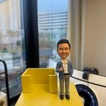 一款為「穩健工程有限公司」主管訂製的人像公仔卡片座，公仔面帶微笑、身穿藍色西裝外套並做出「讚好」手勢，站在一個鮮黃色的實用卡片座旁，是一款充滿正能量的工程師禮物或升職禮物。