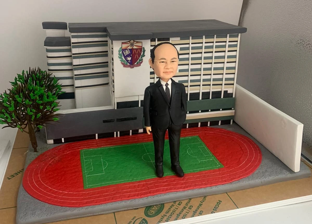 一位身穿西裝的校長的人像公仔,莊重地站立在他服務的學校建築模型前,模型旁有紅色的運動跑道和一個小樹,這是一款向教育工作者致敬的榮休禮物。