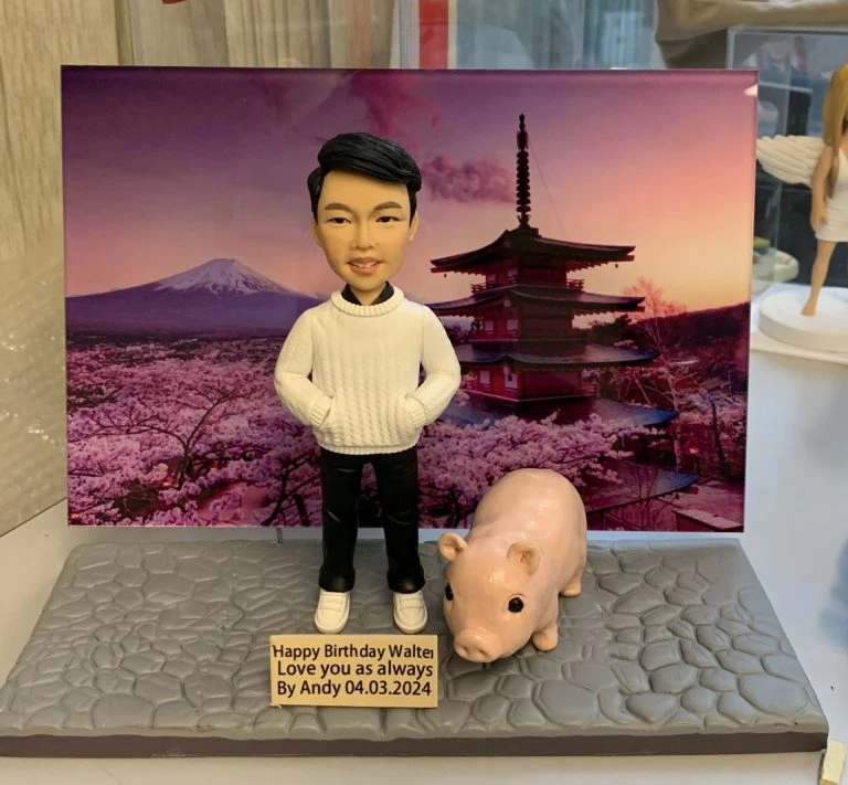 一個穿著白色毛衣的人像公仔站在石板地上，旁邊有一隻小豬模型，背景是櫻花和日本寺廟，底座上有英文生日祝福字牌。