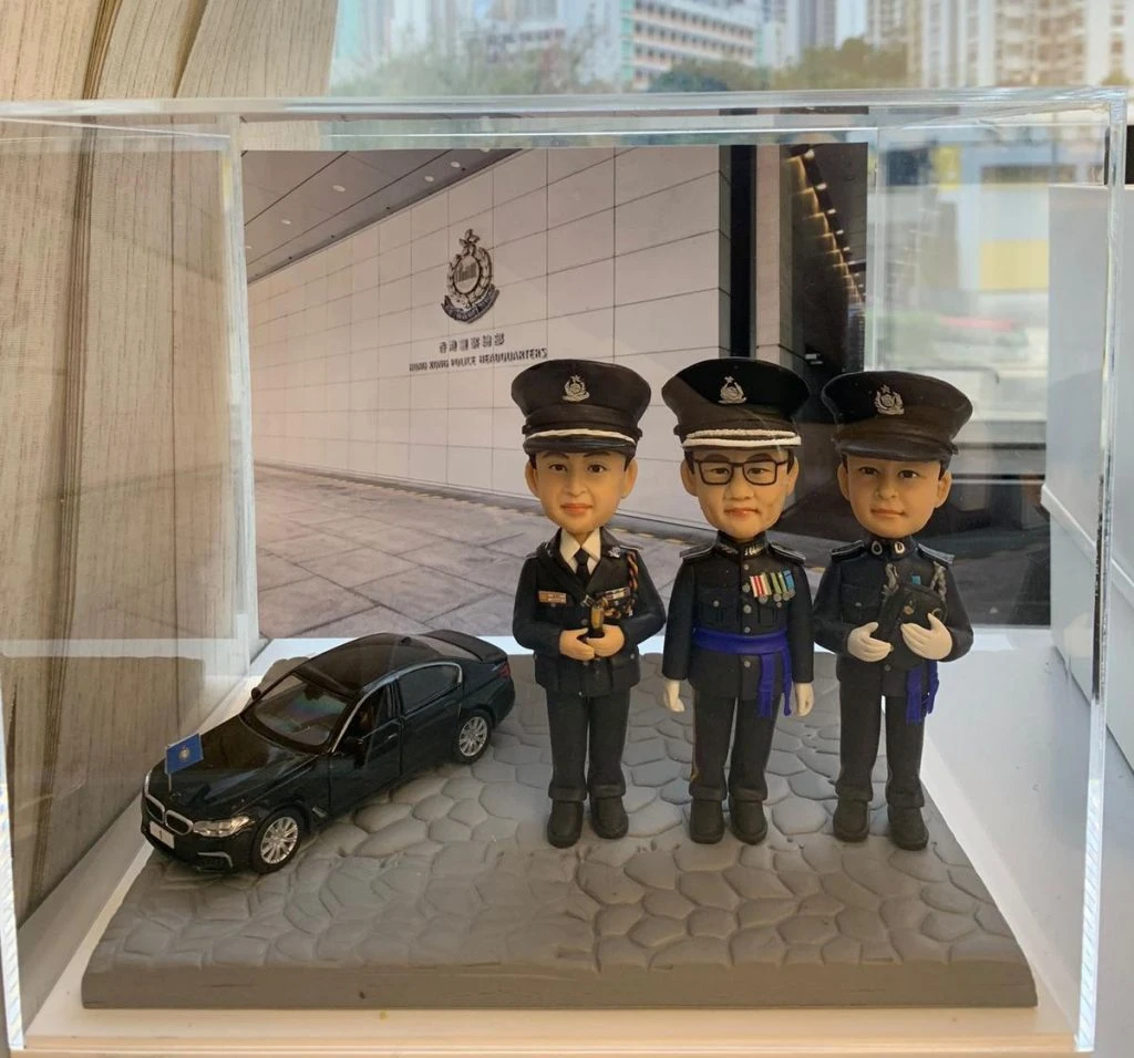 一組包含三位香港警務人員的人像公仔，他們身穿莊嚴的警隊禮服，站在香港警察總部背景前。中間的指揮官佩戴著勳章，旁邊還有一輛專車模型，是一份極具份量的團隊榮休禮物或歡送紀念品。