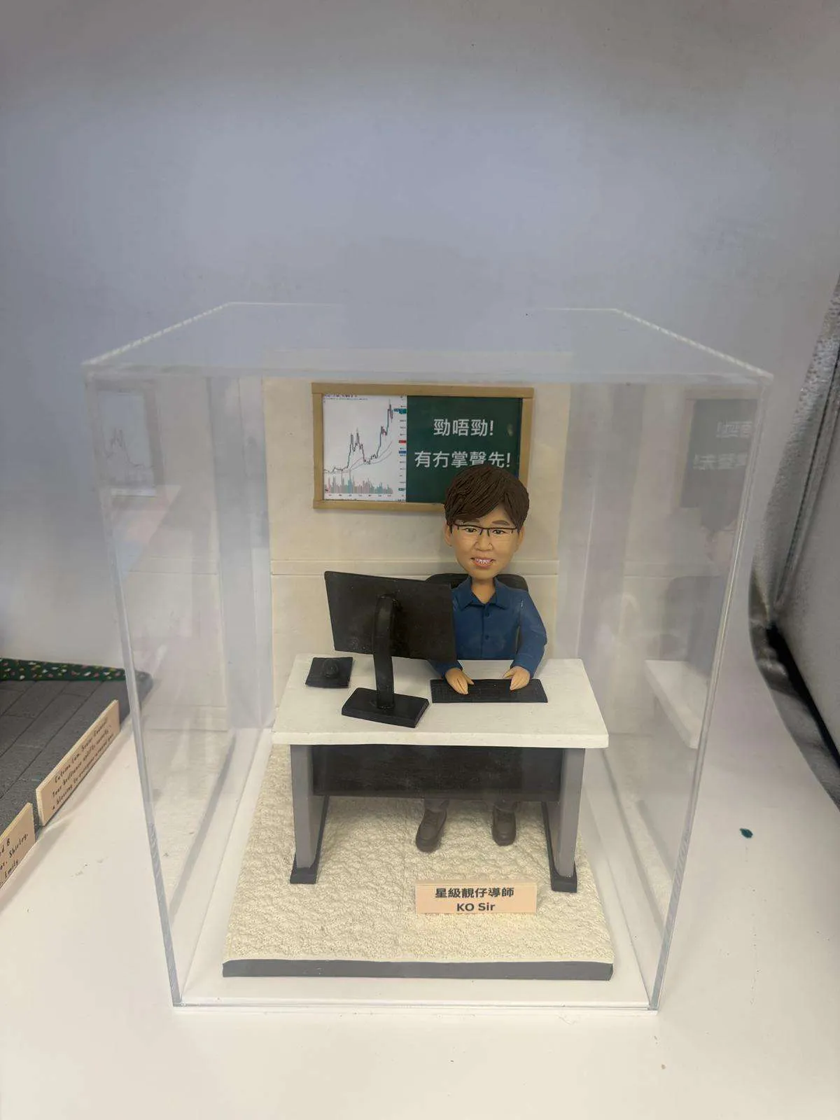 Q版真人公仔KO Sir身穿藍色襯衫坐在辦公桌前，桌面有電腦和鍵盤，背景是寫有「勤唔勤！有冇掌聲先！」的股票圖表黑板，是訂造人像公仔送給教育權威KO Sir的專屬禮物。
