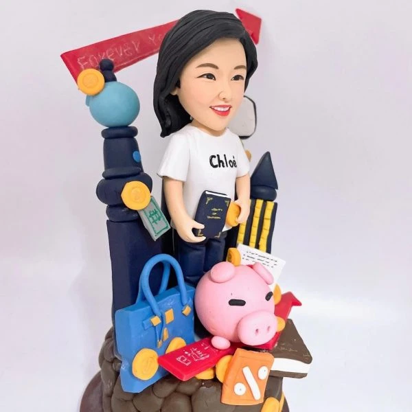 一座以理財與旅遊為主題的人像公仔：黑髮女子穿白色短袖T恤與黑裙，手持深藍色護照與金幣，站在滿載物件的場景前。周圍擺有藍色手提包、信用卡、粉紅小豬撲滿、金幣、紅色百分比折扣牌、書本、收據與計算器意象；背景有黑色行李箱、柱狀與箭頭上升的圖表，以及手機螢幕與鉛筆組，象徵投資成長與工作效率。整體以紅、藍、金為主色，底座寫著品牌字樣，呈現活潑俏皮的財務規劃與旅行熱愛主題。