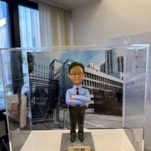 一尊以「警長」職級為主題的警察人像公仔，置於透明壓克力展示盒中，前景由毛絨袖口的手掌托起。公仔短黑髮、戴黑框眼鏡、露齒微笑；身穿淺藍色長袖制服襯衫，肩上有黑色肩章與銀色扣件，領口繫黑色領帶，袖口整齊收邊；下身為深色長褲與黑色皮鞋。公仔雙臂在胸前交叉，姿態沉著自信，站立於灰色六角石紋台座上。台座前方固定金色名牌，刻有祝賀與年份字樣。展示盒後插入的背景板為一座警署建築外觀與鐵欄圍欄的街景，遠處可見高樓大廈與藍天白雲。展示盒外仍能看見室內辦公環境元素，如落地窗邊框、百葉窗簾與牆面，左側窗面反射出長條燈光。整體畫面營造出在職場場景中為「警署警長榮升」而準備的紀念與送禮氛圍。