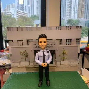 作品以立正合掌的警察人像公仔為主角，透過手作雕刻強調臉部比例、眉眼刻線與唇型弧度，短髮髮束以細筆分層上色呈現光澤；制服部分以淺藍襯衫配黑色領帶與長褲，口袋蓋、鈕扣、肩章和袖口褶位皆以刻紋與陰影處理表現層次，胸前識別章樣式徽片與肩掛對講機組件增添實務感。地台採顆粒化肌理，背板直出警署正門影像，構成「在崗位前敬禮」的情境，既具職務辨識也富儀式感。成品適合作為榮升、退休或嘉許場合的體面贈禮與辦公室展示，亦可客製姓名、職級、頒贈日期與隊章元素，或替換為所屬單位建築背板，使作品更貼近主角故事。