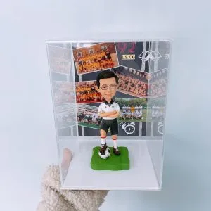 足球運動人像公仔,身穿白色球衣與黑色短褲,雙手交叉自信站立在足球上,擺放於透明展示盒內,背景為球隊合照。適合作為運動紀念、足球愛好者專屬收藏與禮物。
