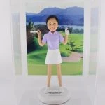 一款客製化人像公仔，呈現一位身穿紫色高爾夫球上衣與白色裙子的女性，肩上揹著高爾夫球桿，臉上帶著笑容。公仔底座寫有「Happy Retirement 2021 from HN Landmark」，背後襯托青山與翠綠草地的高爾夫球場場景，象徵運動、高爾夫球與專屬禮物。
