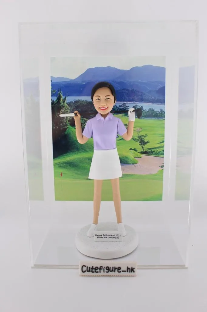 一款客製化人像公仔，呈現一位身穿紫色高爾夫球上衣與白色裙子的女性，肩上揹著高爾夫球桿，臉上帶著笑容。公仔底座寫有「Happy Retirement 2021 from HN Landmark」，背後襯托青山與翠綠草地的高爾夫球場場景，象徵運動、高爾夫球與專屬禮物。