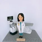 女性醫生人像公仔，身穿白袍與藍綠色醫護制服，脖子掛著聽診器，站在診症室中。背景配置超聲波（Ultrasound）檢查儀與螢幕，上方顯示胎兒影像，旁邊有檢查床，完整呈現婦產科或內科檢查場景。此專屬客製公仔適合作為醫生禮物、醫護人員升職賀禮、退休紀念或醫療團隊感謝禮物。