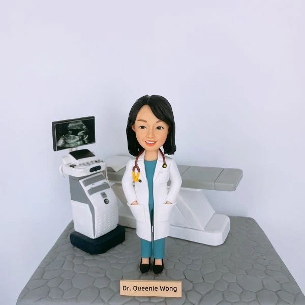 女性醫生人像公仔，身穿白袍與藍綠色醫護制服，脖子掛著聽診器，站在診症室中。背景配置超聲波（Ultrasound）檢查儀與螢幕，上方顯示胎兒影像，旁邊有檢查床，完整呈現婦產科或內科檢查場景。此專屬客製公仔適合作為醫生禮物、醫護人員升職賀禮、退休紀念或醫療團隊感謝禮物。