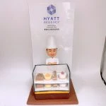 酒店甜品主題的人像公仔擺設：穿白色主廚服與高帽的酒店甜品師傅，雙手抱胸微笑站在玻璃甜品櫃後。甜品櫃內分兩層陳列迷你蛋糕與糕點，包括長條蛋糕、慕斯圈、裝飾莓果的小蛋糕等；底座為棕色方形。背板印有「HYATT REGENCY HONG KONG TSIM SHA TSUI 香港尖沙咀凱悅酒店」標誌與字樣。整體風格典雅，適合作為酒店甜品師傅退休禮物與紀念擺設。