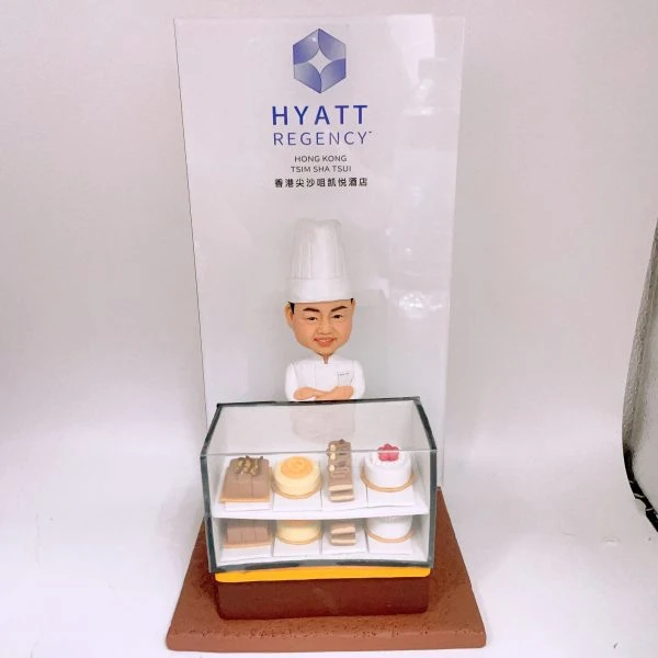 酒店甜品主題的人像公仔擺設：穿白色主廚服與高帽的酒店甜品師傅，雙手抱胸微笑站在玻璃甜品櫃後。甜品櫃內分兩層陳列迷你蛋糕與糕點，包括長條蛋糕、慕斯圈、裝飾莓果的小蛋糕等；底座為棕色方形。背板印有「HYATT REGENCY HONG KONG TSIM SHA TSUI 香港尖沙咀凱悅酒店」標誌與字樣。整體風格典雅，適合作為酒店甜品師傅退休禮物與紀念擺設。