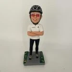 一款客製化人像公仔，公仔戴著黑色單車安全帽與紅色運動墨鏡，身穿白色運動上衣與黑色緊身單車褲，雙手交叉胸前，自信站立於灰色石磚道路上，兩側綠色草地點綴著小花，展現熱愛單車運動的個人特色與活力氛圍。