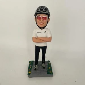 一款客製化人像公仔，公仔戴著黑色單車安全帽與紅色運動墨鏡，身穿白色運動上衣與黑色緊身單車褲，雙手交叉胸前，自信站立於灰色石磚道路上，兩側綠色草地點綴著小花，展現熱愛單車運動的個人特色與活力氛圍。