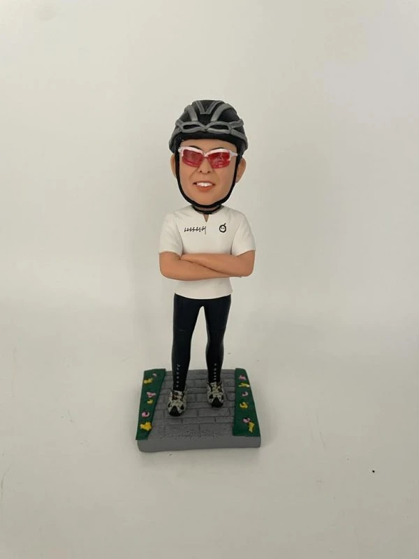 一款客製化人像公仔，公仔戴著黑色單車安全帽與紅色運動墨鏡，身穿白色運動上衣與黑色緊身單車褲，雙手交叉胸前，自信站立於灰色石磚道路上，兩側綠色草地點綴著小花，展現熱愛單車運動的個人特色與活力氛圍。