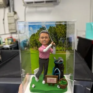 【老闆退休】哥爾夫球主題人像公仔:中年男士公仔穿粉紅條紋高爾夫上衣、深灰長褲與白球鞋,雙手完成揮桿動作站在綠色草地底座上;身後有黑藍相間的球桿袋與多支球桿,腳邊散落白色高爾夫球,前方木牌寫有祝賀與日期字樣。背景為樹林環繞的球道照片板;整體置於透明防塵盒內,呈現「老闆退休」慶賀與休閒品味的哥爾夫球人像公仔。