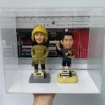 一款為消防員訂製的退休人像公仔禮物，包含兩個公仔，一個穿消防制服，另一個穿休閒服，背景是上環消防局照片，裝在透明保護盒中。