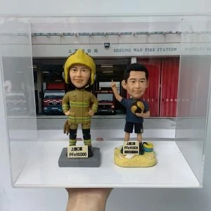 一款為消防員訂製的退休人像公仔禮物，包含兩個公仔，一個穿消防制服，另一個穿休閒服，背景是上環消防局照片，裝在透明保護盒中。